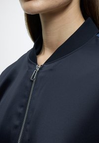 Μπουφάν βομβαρδιστικού ναυτικού μπλε με κλείσιμο με φερμουάρ, ριμπ κολάρο και λεία υφή υφάσματος. Κοντινή λήψη που τονίζει τα σχέδια και τις λεπτομέρειες του υλικού.