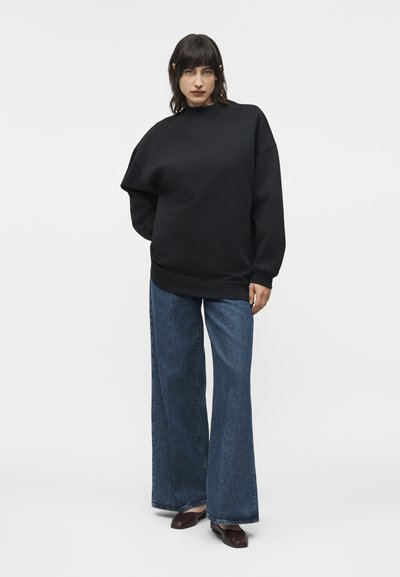 Γυναίκα που στέκεται φορώντας φαρδιά μαύρη μπλούζα, φαρδιά μπλε τζιν παντελόνια και καφέ επίπεδα παπούτσια, μπροστά σε απλό ανοιχτό φόντο.