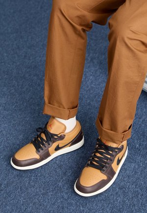 Braune und beige Leder-Sneaker mit perforierter Spitze, schwarzen Schnürsenkeln und roten Akzenten; kombiniert mit hochgekrempelten braunen Hosen, auf einem blauen Teppich platziert.