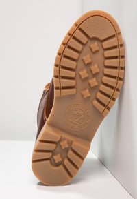 Suela de bota de goma marrón con un patrón texturizado, que presenta un logotipo circular y un número de modelo, mostrada desde una perspectiva inferior.