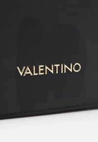 Un material negro texturizado presenta un prominente logo dorado "VALENTINO", con un sutil patrón de fondo y bordes cosidos.