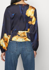 Fließende schwarze Bluse mit großen gelben Blumenmustern, runder Ausschnitt und langen Ärmeln, kombiniert mit hoch taillierten blauen Jeans.