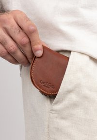 Braune Lederbörse mit abgerundeten Kanten, genähten Details und einem geprägten Logo, teilweise in einer hellfarbenen Stofftasche gehalten.