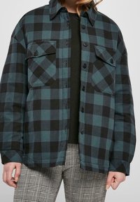 Camisa de franela en cuadros de azul oscuro y negro. Presenta dos bolsillos en el pecho, cierre de botones y un corte relajado. Lleva una camiseta negra debajo.