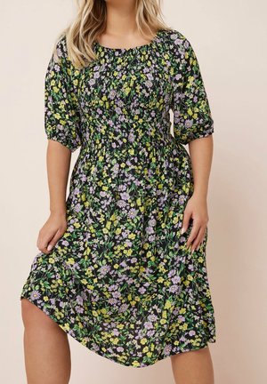 Kobieta w czarnej sukience midi z żółto-fioletowym kwiatowym wzorem, bufkami i marszczonym stanem na jednolitym beżowym tle.