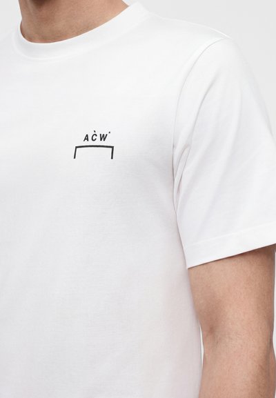 Λευκό t-shirt με κοντά μανίκια και μικρό μαύρο λογότυπο "A C W *" πάνω από μια μαύρη οριζόντια γραμμή στο αριστερό στήθος, φορεμένο από ένα άτομο.