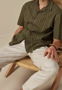 Camisa corta de manga corta en verde oliva con patrones texturizados y cierre de botones, combinada con pantalones beige claro, sentado en una silla de mimbre.