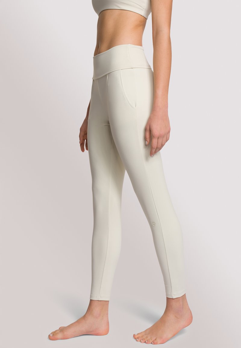 Leggings high-waisted color beige chiaro realizzati in tessuto elasticizzato, con tasche laterali, una vestibilità aderente e una texture liscia. Logo vicino alla caviglia.