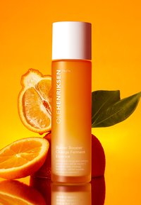 Orangene Flüssigkeit in einer transparenten Flasche mit einem weißen Deckel, etikettiert mit "Barrier Booster Orange Ferment Essence." Orangenscheiben und Blätter in der Nähe.