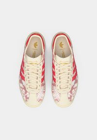 Adidas-sneakers med et rødt og hvitt blomstermønster, kremfarget mesh-overdel, tre røde striper, og gull-logo detaljer på tungen og sålen.