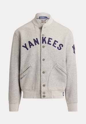 Zwei Models tragen graue Yankees-Jacken und Jeans, eines mit weißen Sneakers, das andere mit blauen Sneakers und einer Yankees-Kappe.