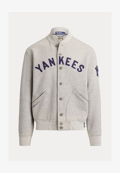 Veste varsity gris clair boutonnée avec "YANKEES" en bleu marine sur le devant, poches inclinées et logo des New York Yankees sur la manche gauche.