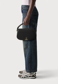 Svart läderhandväska med en strukturerad form, böjd handtag och guldfärgad detaljer; modell som bär väskan tillsammans med avslappnade blå denimjeans och sneakers.