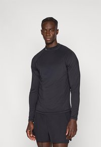 Hosszú ujjú fekete sportfelső sima, rugalmas anyagból, testhezálló kivitelben, diszkrét logó a gallérnál, fekete rövidnadrággal párosítva.