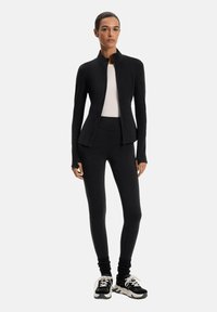 Giacca sportiva nera con colletto alto, design aderenza e maniche lunghe, abbinata a leggins neri. Modella con sneakers nere.