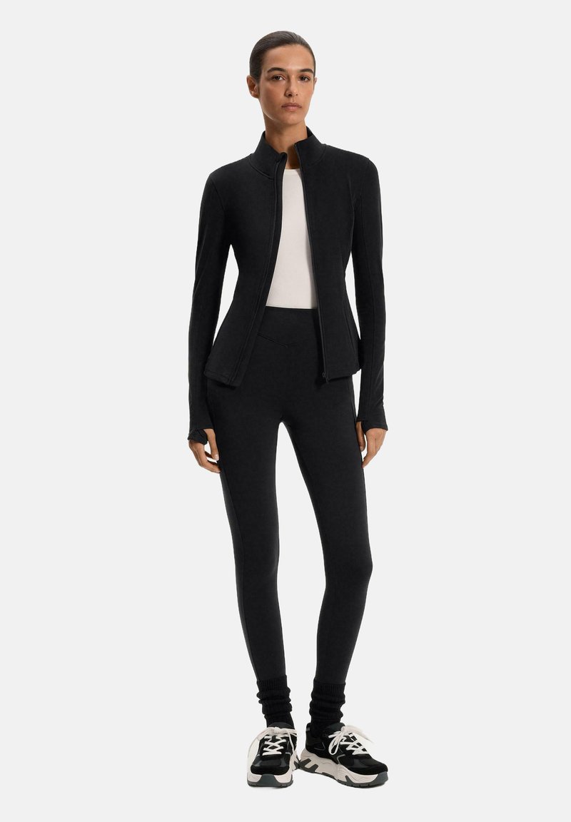 Giacca sportiva nera con colletto alto, design aderenza e maniche lunghe, abbinata a leggins neri. Modella con sneakers nere.