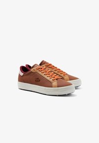 Bruna lädersneakers med orange snören, vita sulor, vinröd innersula och en krokodil-logotyp på sidan, visade i ett par vända till höger.