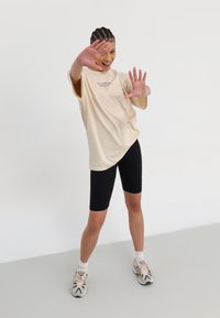 Beige oversized t-skjorte med logo, sorte sykkelskjørt, og flerfargede treningssko. Motivet poserer med utstrakte hender.