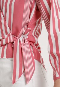 Blusa a righe rosa e bianche con maniche lunghe, caratterizzata da un dettaglio nodo alla vita. Realizzata in un tessuto leggero e morbido.