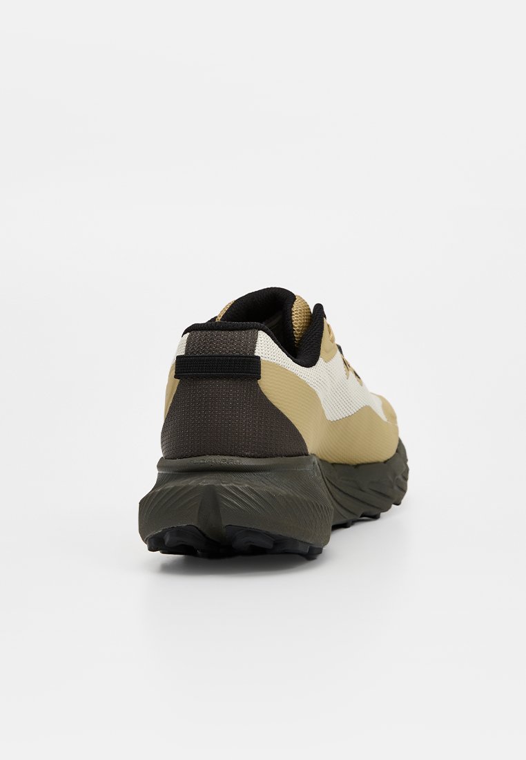Zapatilla deportiva beige y blanca con detalles en negro, suela gruesa y texturizada, y lengüeta para tirar del talón, mostrada desde el ángulo trasero sobre un fondo liso.