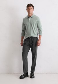 Cămașă polo verde deschis cu mânecă lungă și guler, combinată cu pantaloni gri închis și mocasini negri, stând în fața unui fundal alb texturat.