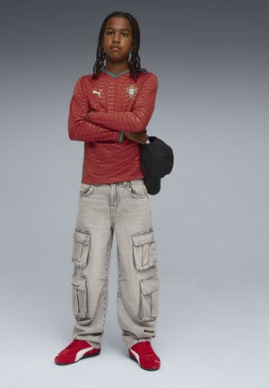 Ung person med flätat hår som bär en röd Portugal-fotbollströja, grå cargobyxor och röda sneakers, håller en svart keps och står med armarna i kors.