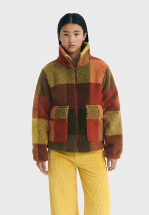 Jeune personne portant une veste sherpa zippée avec de grandes poches à rabat dans un motif écossais rouge, orange et vert, associée à un pantalon en velours côtelé jaune.