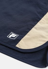 Tissu bleu marine et beige avec un logo blanc représentant la lettre « F » stylisée près de l'ourlet d'un vêtement, probablement un short ou un pantalon.