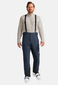 Marineblaue wasserdichte Schneehose mit verstellbaren Hosenträgern, seitlichen Reißverschlusstaschen und verstärkten unteren Beinen. Kombiniert mit einem hellgrauen Langarmshirt.