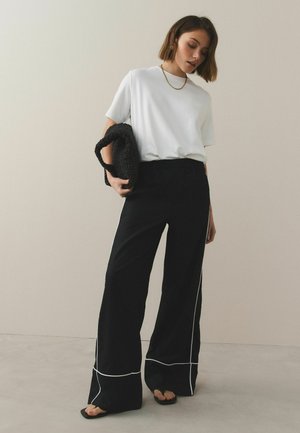Frau trägt weißes kurzärmeliges Oberteil und schwarze, weit geschnittene Hose mit weißer Paspelierung, hält eine schwarze, strukturierte Handtasche und steht auf hellem Boden.