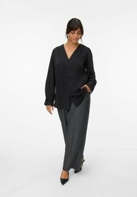 Blouse en satin noir avec un décolleté en V, des manches longues et une fermeture à boutons. Associée à un pantalon gris à jambes larges et des escarpins noirs à talon pointu.