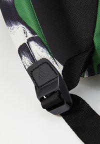 Black adjustable strap with textured material and a black plastic clasp featuring a logo. Green and patterned fabric background.