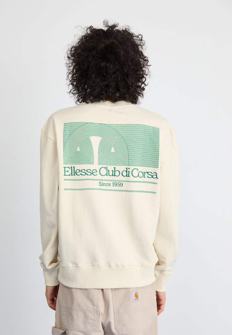Krémový svetr s zeleným grafickým designem na zadní straně, který obsahuje vlnité čáry a text "Ellesse Club di Corsa Since 1959."