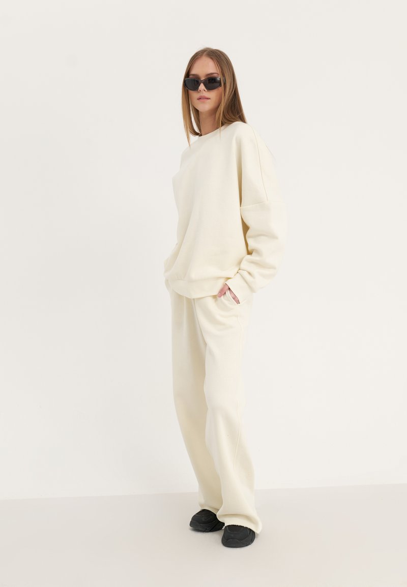 Sweatshirt oversize beige chiaro abbinato a pantaloni coordinati, con una vestibilità comoda e tessuto morbido. Occhiali da sole neri completano il look.