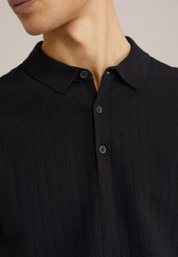 Polo shirt tricotat negru cu o textură ribbed, având un guler și patru nasturi negri. Vedere de aproape care pune în evidență decolteul.