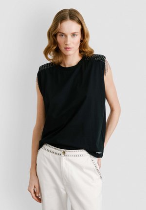 Maglietta nera senza maniche con dettagli di borchie sulle spalle, indossata con pantaloni bianchi con hardware decorativo lungo il girovita.