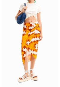 Oranssi ja valkoinen siteellä värjätty huivikrotsi, jossa on solk dettag, yhdistettynä lyhythihaiseen valkoiseen puseroon ja siniseen minikassiin; malli käyttää hihnakenkiä.