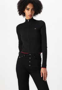 Svart ribbestrikket genser med glidelås og høy krage, med røde detaljer ved kanten, paret med høyt liv svarte jeans med knappelukking.
