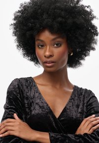 Robe en velours noir avec un décolleté en V profond et des manches longues. Le mannequin a une coiffure afro et pose les bras croisés. Texture lisse et brillante.