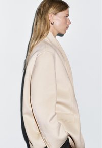 Beige oversized blazer gemaakt van zachte stof met een op maat gemaakte kraag, enkele knoopsluiting en zijzakken. Gladde textuur, minimalistisch design.