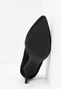 Chaussure à talon haut en daim noir avec un bout pointu et un talon fin. La semelle est lisse et noire, avec des détails de marquage minimaux.