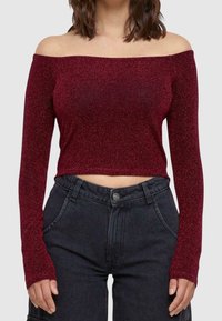 Maroon off-the-shoulder crop top dengan lengan panjang, terbuat dari kain berkilau; dipasangkan dengan celana denim gelap berpinggang tinggi.