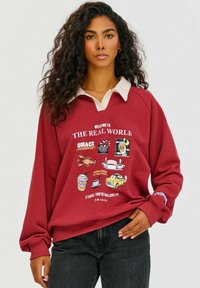 Červená mikina s krémovým límcem, zdobená hravými grafickými motivy a textem na přední straně. Volný střih s dlouhými rukávy, vyrobená z jemné tkaniny.