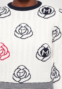 Strikkegenser i hvitt med marineblå kabelmønstre og rosemotiver i marineblått og rødt. Ribbet marineblå halslinning og stripete bunnsøm.