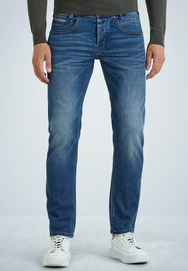 Vaqueros de mezclilla azul desgastados y ajustados, con cinco bolsillos, trabillas para el cinturón y detalles de costura sutiles. Combinados con zapatillas blancas sobre un fondo claro.