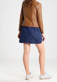 Femme portant une veste en cuir marron, une jupe en denim bleue et des baskets blanches, se tenant de dos.