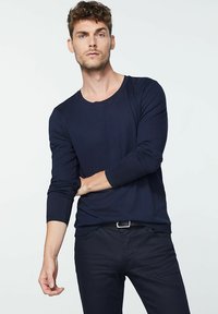 Dunkelblaues Langarm-T-Shirt aus weichem Stoff mit Rundhalsausschnitt. Das Modell posiert mit übergeschlagenem Arm und trägt dunkel enge Hosen.