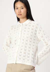 Bela pletena jopica z vzorcem čipke, gumb v sprednji del in rebrast ovratnik ter rob. Dolgi rokavi s sproščenim krojem.