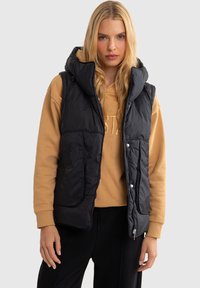 Musthave must vest, milletaskourtes, supergiande jaot kõik ruudud ja käepidemete kaudu, beežhuviti peal, neutraalses taustas.