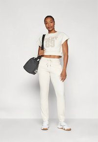 Γυναίκα που στέκεται φορώντας κρεμ μπλούζα crop top με το "DKNY 89", ταιριαστά παντελόνια φόρμας, λευκά αθλητικά παπούτσια, και κρατώντας μια μαύρη τσάντα ώμου σε ανοιχτό φόντο.